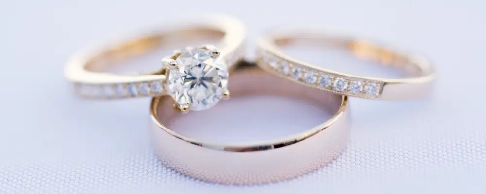 A complete three-ring wedding set in rose gold featuring a round solitaire engagement ring, a diamond-encrusted wedding band, and a matching smooth men’s wedding band.