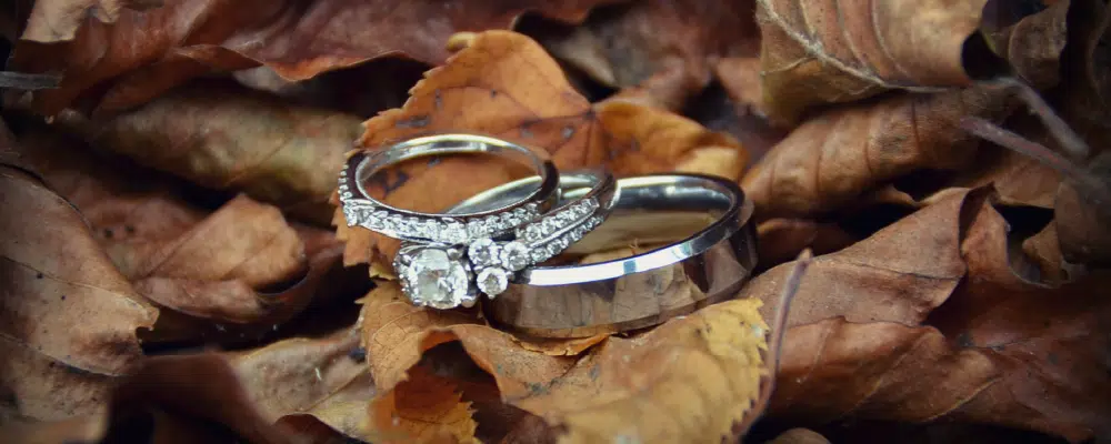 A matching white gold bridal set featuring a diamond engagement ring and wedding band displayed next to a thick men’s platinum wedding ring on fall leaves.