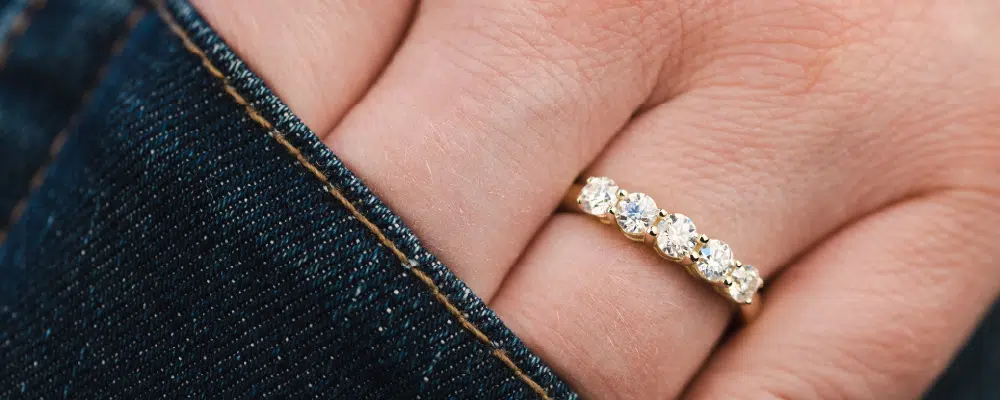 A classic yellow gold 5-stone diamond wedding band worn on a finger next to a denim pocket