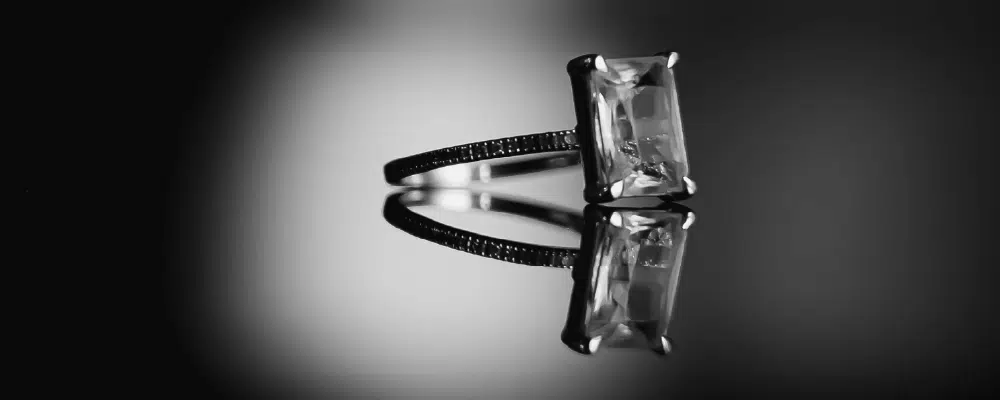 Emerald cut diamond engagement ring in a four-prong setting with a pavé diamond band on a reflective black surface.