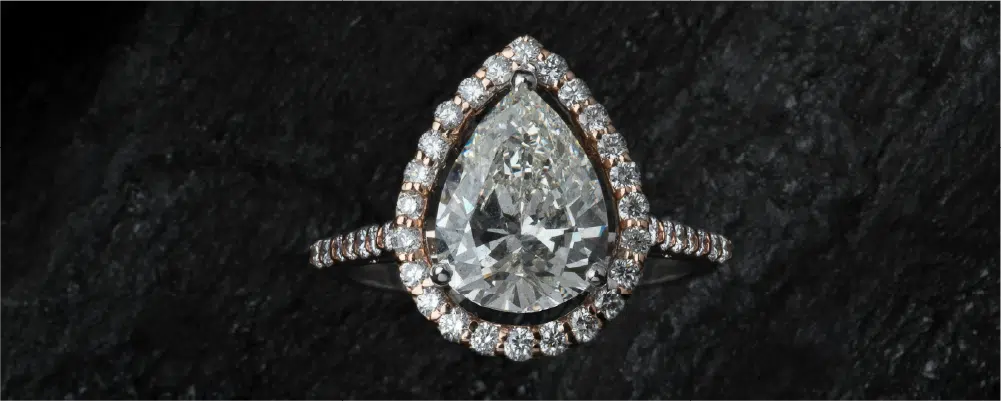 Top-down view of a brilliant pear shaped diamond engagement ring in a halo setting resting on a textured dark stone background.