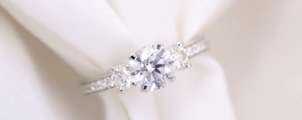 Classic round brilliant three stone engagement ring with a pavé diamond band and white fabric backdrop.