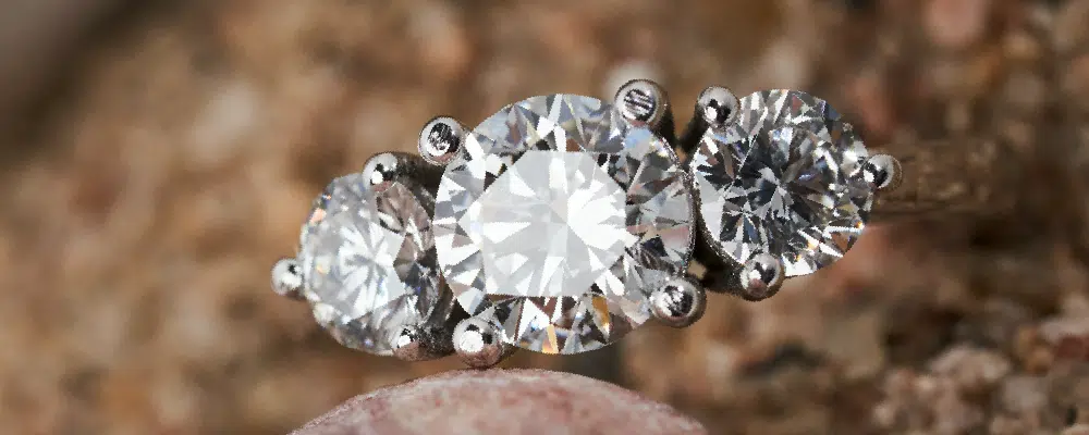 Macro shot of a trilogy engagement ring with three round diamonds in a shared prong setting on a natural stone surface.