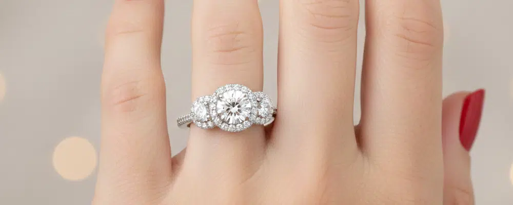 Close up of a classic three-stone round brilliant diamond engagement ring featuring micro-pavé halo borders on a finger