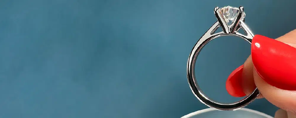 A close-up side profile of a sparkling cathedral solitaire engagement ring held against a blue background, displaying its distinct architectural design.