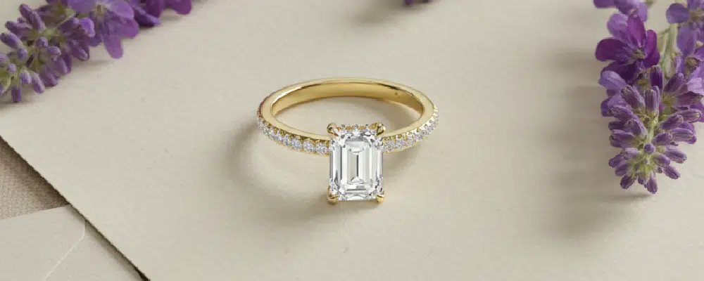 A classic yellow gold emerald cut engagement ring with a delicate diamond pavé band styled next to purple lavender flowers