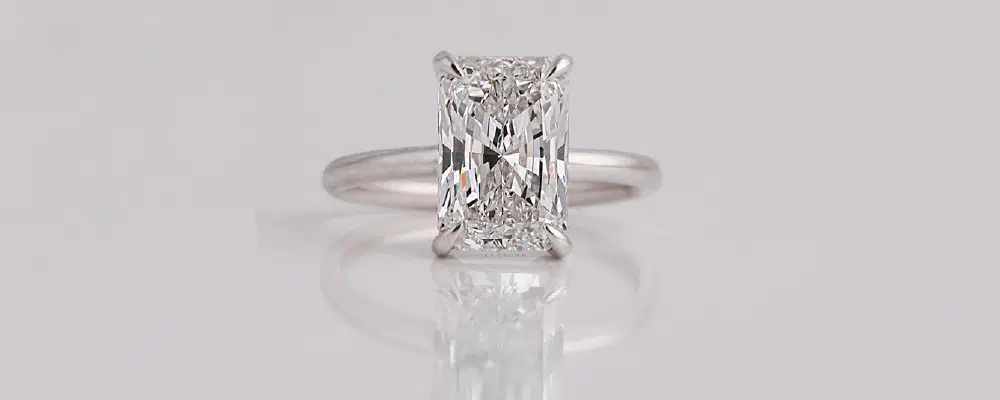 A classic platinum radiant cut solitaire engagement ring with a simple plain band standing on a reflective surface