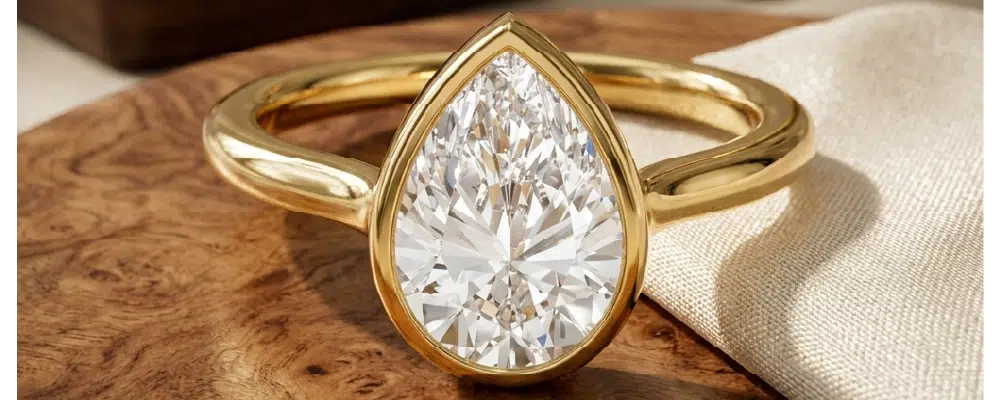 A modern yellow gold bezel set pear engagement ring with a thick protective metal rim on a natural wood background