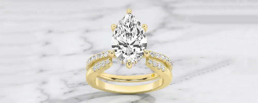 A unique yellow gold pear engagement ring setting featuring a split shank pavé band on a white marble background