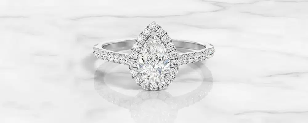 A traditional white gold halo pear engagement ring on a marble surface comparing classic rim halos with a modern hidden halo layout