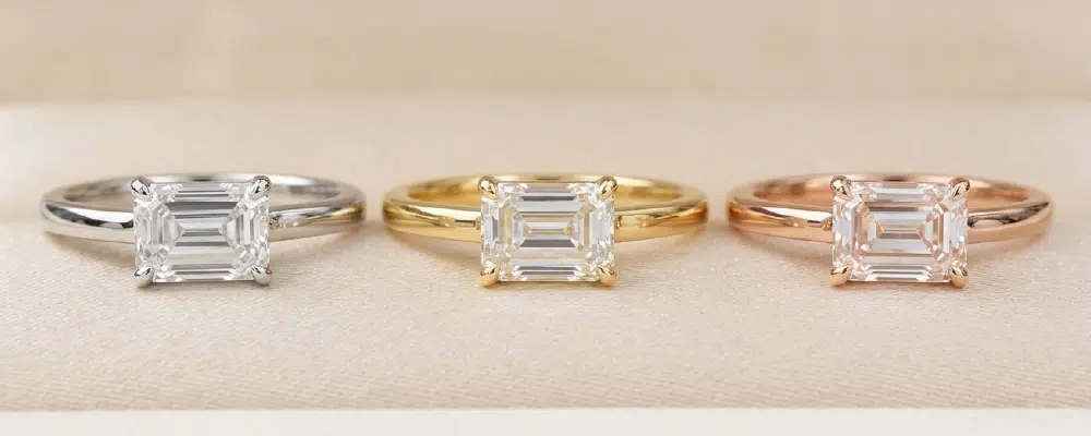 Three east-west emerald cut solitaire engagement rings displayed in white gold yellow gold and rose gold bands