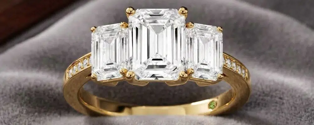 A classic yellow gold 3 stone emerald cut engagement ring with perfectly matched side diamonds and a pave accented band on grey velvet