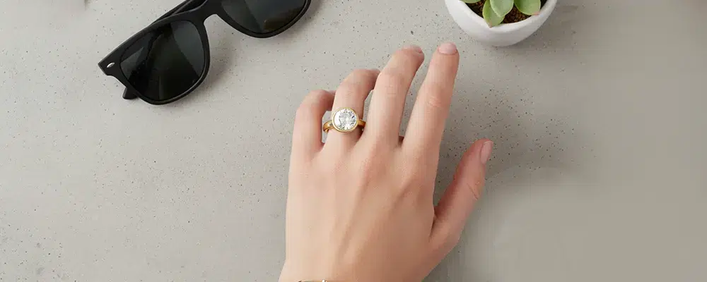 Top-down lifestyle view of a woman wearing a modern bezel set round diamond ring on a neutral stone surface next to sunglasses.