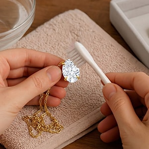 A person's hands holding a yellow gold pear-cut lab-grown diamond pendant necklace over a brown textured towel, gently cleaning it with a white toothbrush, with a small glass bowl of water resting in the background.