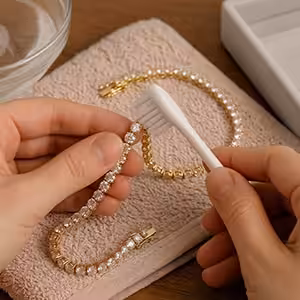A person's hands holding a yellow gold round-cut lab-grown diamond tennis bracelet over a brown textured towel, gently cleaning it with a white toothbrush, with a small glass bowl of water resting in the background.
