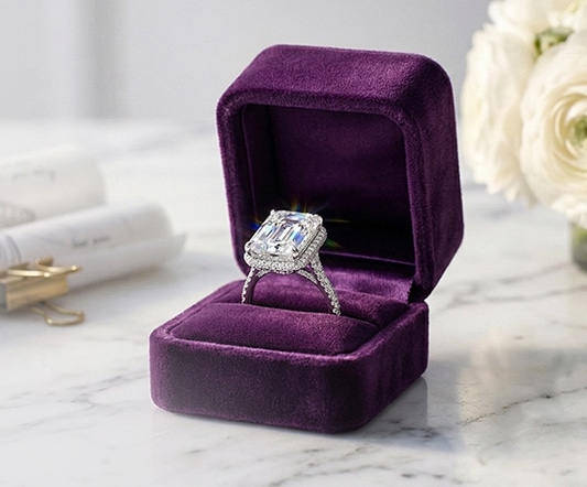 Custom-designed emerald-cut lab-grown diamond engagement ring presented in a purple velvet jewelry box next to a white rose.