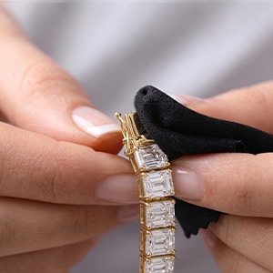 A close-up of hands using a black microfiber cloth to carefully hold and inspect the clasp of a thick yellow gold emerald-cut lab-grown diamond tennis bracelet.