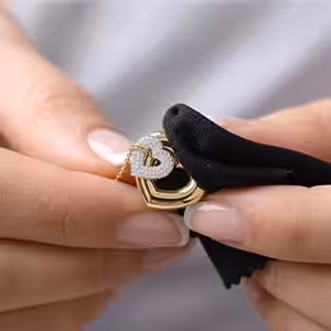 A close-up of hands using a black microfiber cloth to carefully hold and inspect a yellow gold necklace featuring an interlocking heart pendant with lab-grown diamond pavé.