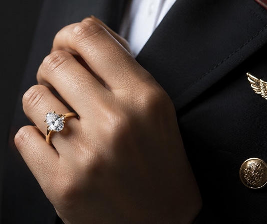 A close-up of a hand wearing a La Joya yellow gold pear-shaped lab-grown diamond engagement ring, resting against a dark uniform jacket featuring gold buttons and a winged insignia, representing the Honorary Program.