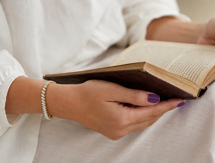 A close-up of a person in a white garment holding an open vintage book, wearing purple nail polish and a La Joya yellow gold lab-grown diamond tennis bracelet, representing the discount program for educators and teachers.