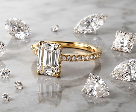 A La Joya yellow gold emerald-cut lab-grown diamond engagement ring with a pavé diamond band, surrounded by a scattered assortment of loose lab-grown diamonds in various shapes—including round, pear, marquise, and princess cuts—resting on a light, polished marble surface, representing heritage craftsmanship and structural integrity.
