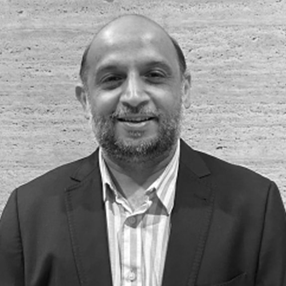 A black and white professional portrait of La Joya President and CEO Nishit Mehta, smiling and wearing a dark suit jacket over a light collared shirt, standing against a light textured wall.