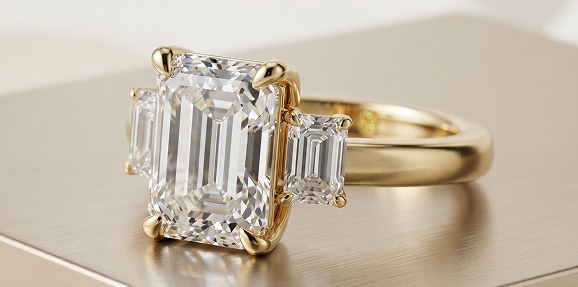 A close-up of a La Joya yellow gold three-stone lab-grown diamond engagement ring featuring a large emerald-cut center stone flanked by two smaller step-cut side stones on a solid gold band, resting on a warm, polished metallic surface.