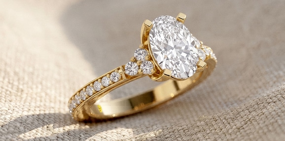 A close-up of a La Joya yellow gold engagement ring featuring an oval-cut lab-grown diamond center stone and a pavé diamond band, resting on a softly lit, textured light beige fabric surface.