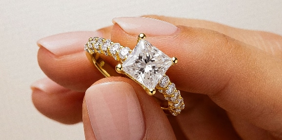 A close-up of a person's fingers delicately holding a La Joya yellow gold lab-grown diamond engagement ring, featuring a prominent princess-cut center stone and a channel-set diamond band against a smooth neutral background.