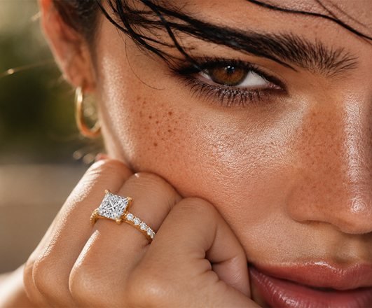Woman resting her hand near her face, wearing a yellow gold princess-cut lab-grown diamond engagement ring and gold hoop earrings.