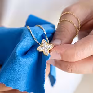 A close-up of hands using a bright blue microfiber cloth to gently hold and polish a yellow gold floral motif lab-grown diamond pendant necklace.