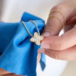 A close-up of hands using a bright blue microfiber cloth to gently hold and polish a yellow gold floral motif lab-grown diamond pendant necklace.
