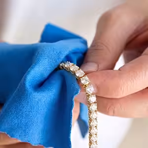 A close-up of hands using a bright blue microfiber cloth to gently hold and wipe a yellow gold round-cut lab-grown diamond tennis bracelet.