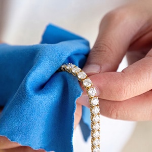 A close-up of hands using a bright blue microfiber cloth to gently hold and wipe a yellow gold round-cut lab-grown diamond tennis bracelet.