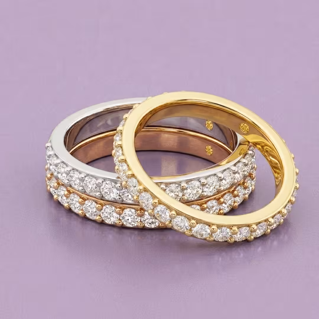 A stack of three round-cut lab-grown diamond eternity rings in white gold, rose gold, and yellow gold on a purple background.
