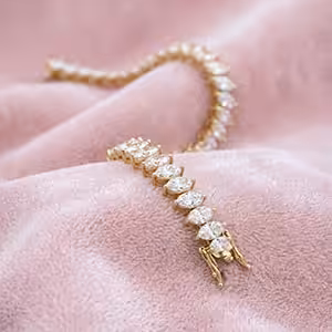A yellow gold pear-cut lab-grown diamond tennis bracelet elegantly draped and resting face-up on a soft, textured pink fabric background.