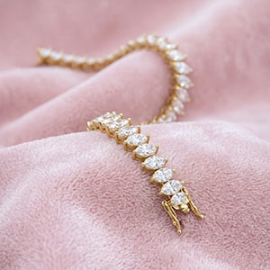 A yellow gold pear-cut lab-grown diamond tennis bracelet elegantly draped and resting face-up on a soft, textured pink fabric background.