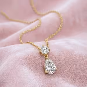 A yellow gold necklace featuring a round-cut and pear-cut lab-grown diamond drop pendant elegantly resting on a soft, textured pink fabric background.