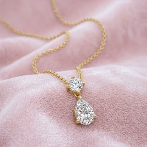 A yellow gold necklace featuring a round-cut and pear-cut lab-grown diamond drop pendant elegantly resting on a soft, textured pink fabric background.
